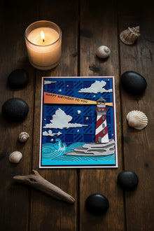 Lighthouse at Night card flat on dark weathered wood with seashells and candlelight